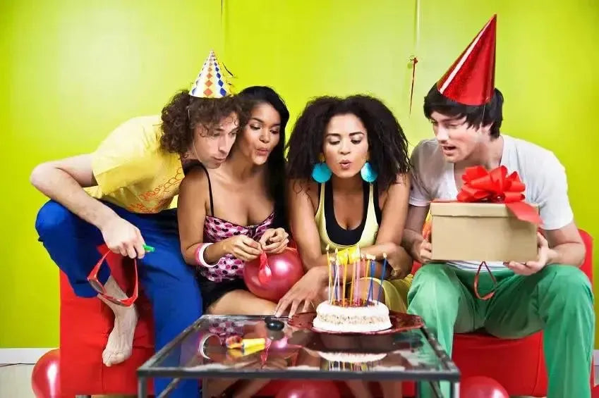 Group of adults at birthday party with hats, cake, candles, and a wrapped gift on a red couch