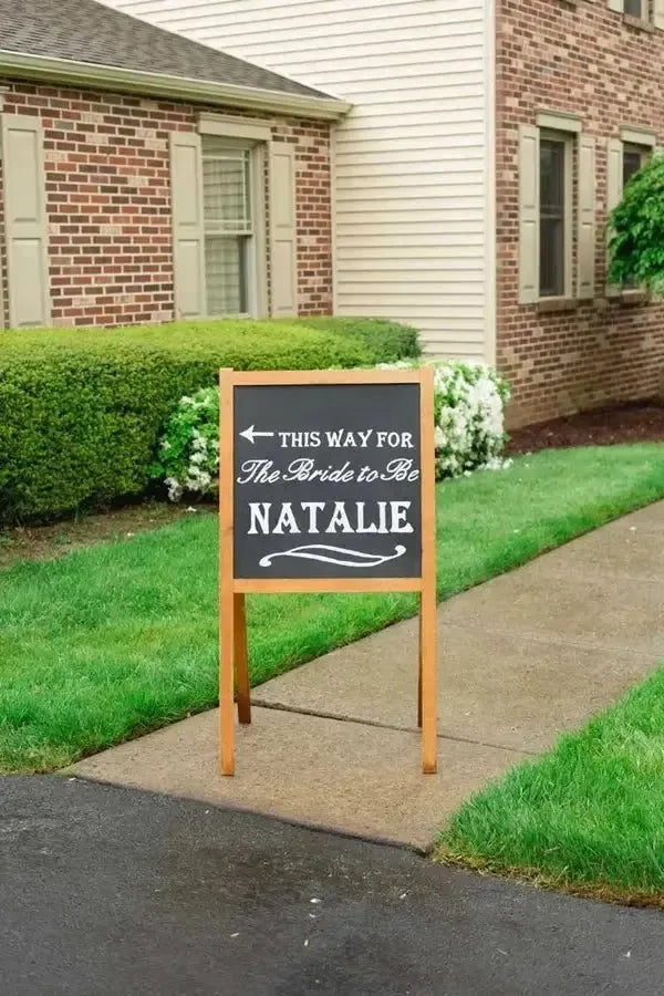 Outdoor bridal shower sign for Natalie on a sidewalk near a brick house and green lawn