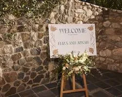 Wedding welcome sign on easel with floral arrangement, stone wall background, outdoor event