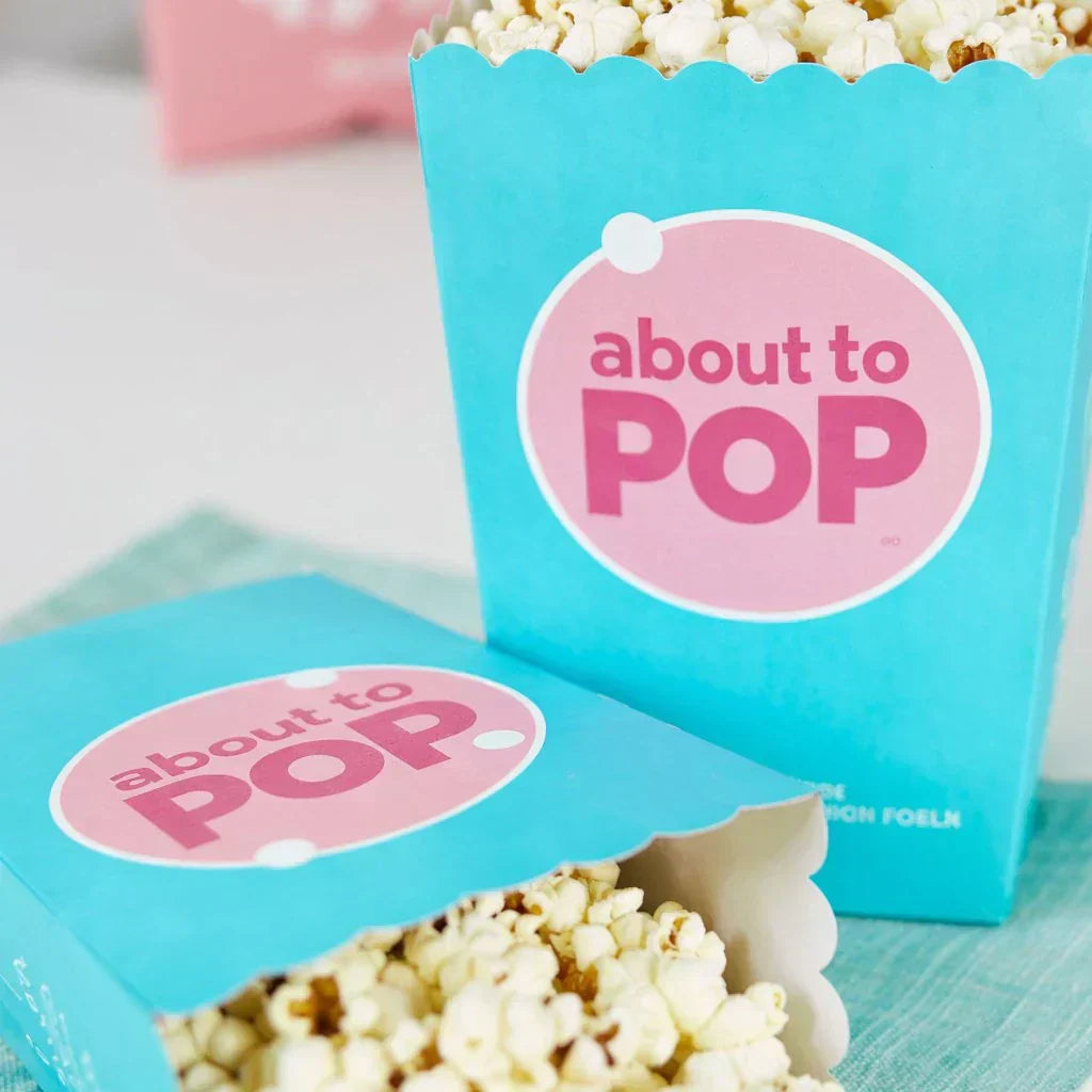 Blue popcorn boxes with pink 'about to pop' baby shower labels, filled with popcorn.