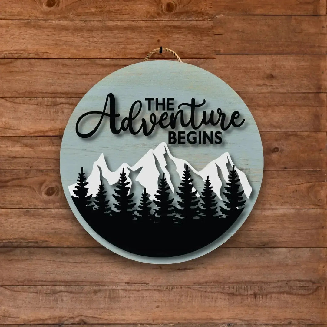 Round custom camping sign with mountain and pine tree design, text 'The Adventure Begins' on wood background.