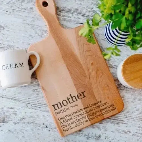 Personalized handle cutting board engraved with mother definition, coffee mug, and plant on table