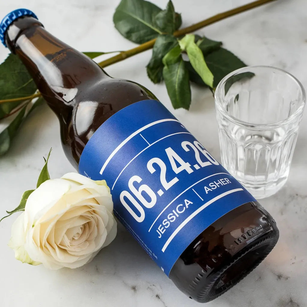 Custom wedding beer bottle with personalized blue label, white rose, and glass on marble table