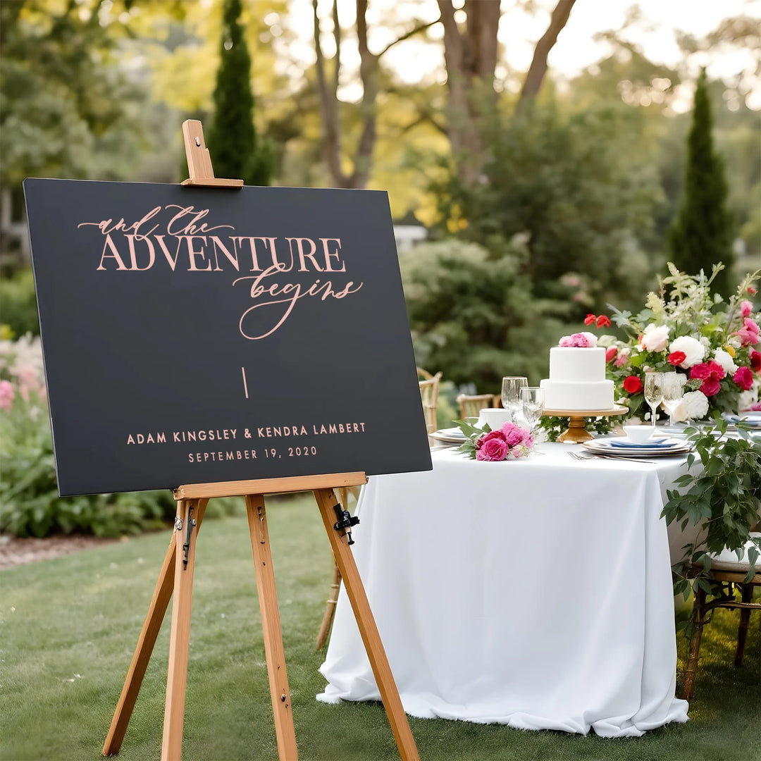 Elegant black wedding welcome sign on easel with pink script next to white cake and floral table outdoors
