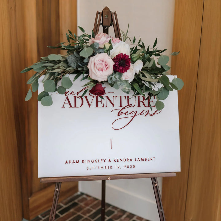 Wedding welcome sign on easel with floral arrangement and text 'Adventure begins' with couple names and date