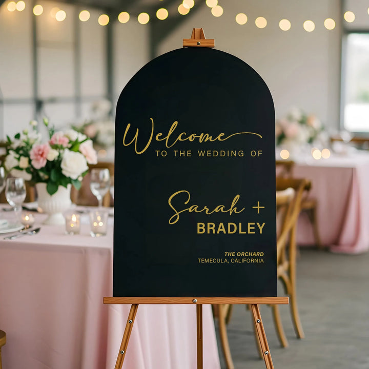 Black arched wedding welcome sign on wooden easel with gold script text at floral decorated reception