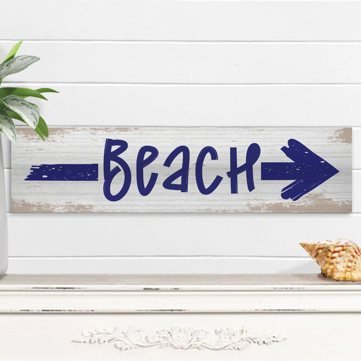 Rustic white wooden beach sign with navy arrow pointing right, placed on shelf with seashell and plant