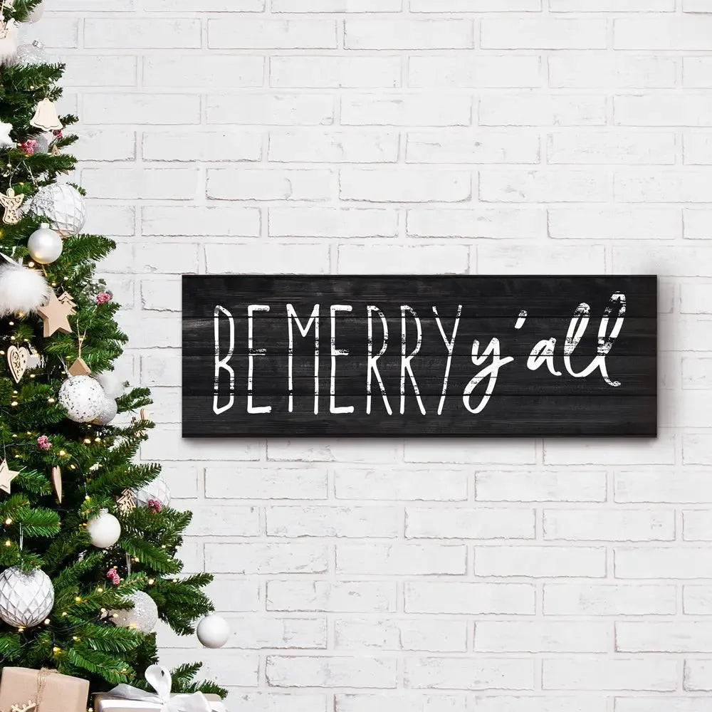Be Merry y'all wood sign on white brick wall beside decorated Christmas tree