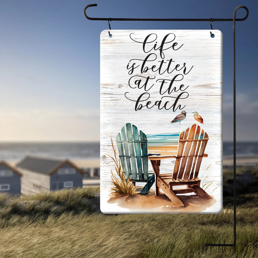Metal garden flag with beach scene and text life is better at the beach, hanging outdoors near grassy dunes