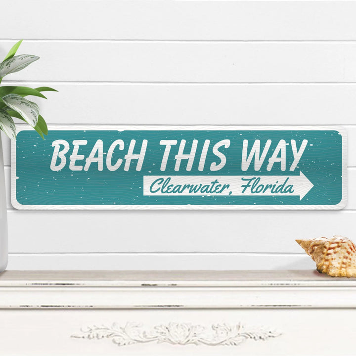 Teal "Beach This Way Clearwater, Florida" metal sign on white wooden wall with plant and seashell