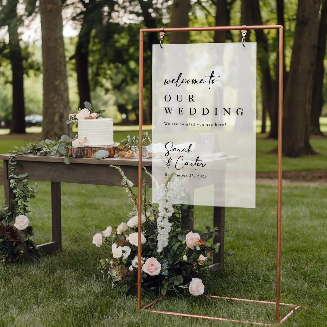 Minimalist acrylic wedding welcome sign with copper frame, floral accents, and white cake on wooden table outdoors