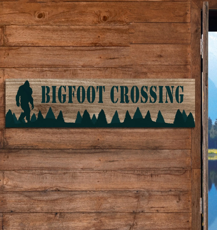 Wooden rustic sign on cabin wall with Bigfoot silhouette, forest trees, and text Bigfoot Crossing