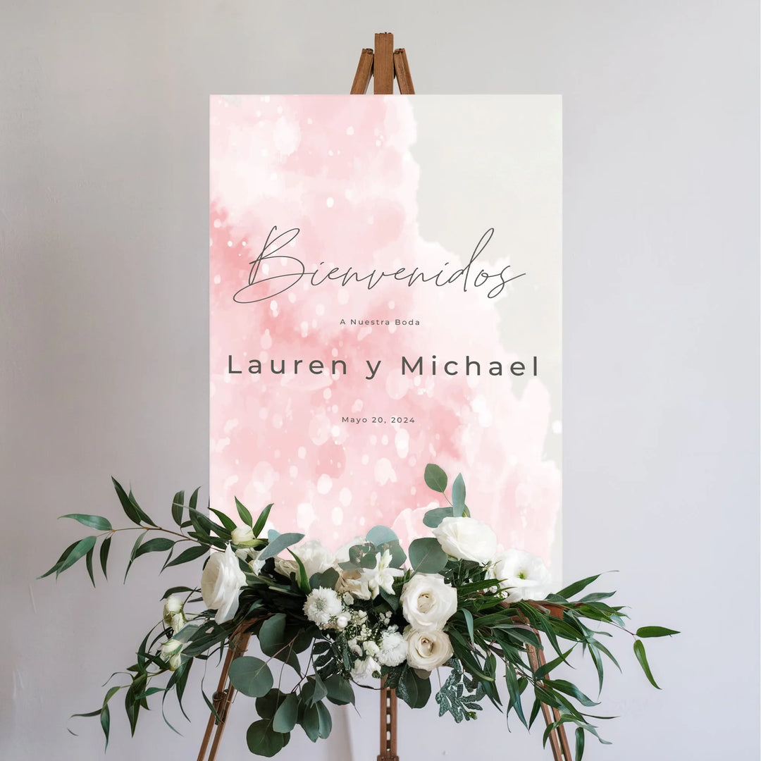 Blush pink Spanish wedding welcome sign with white flowers and greenery on wooden easel
