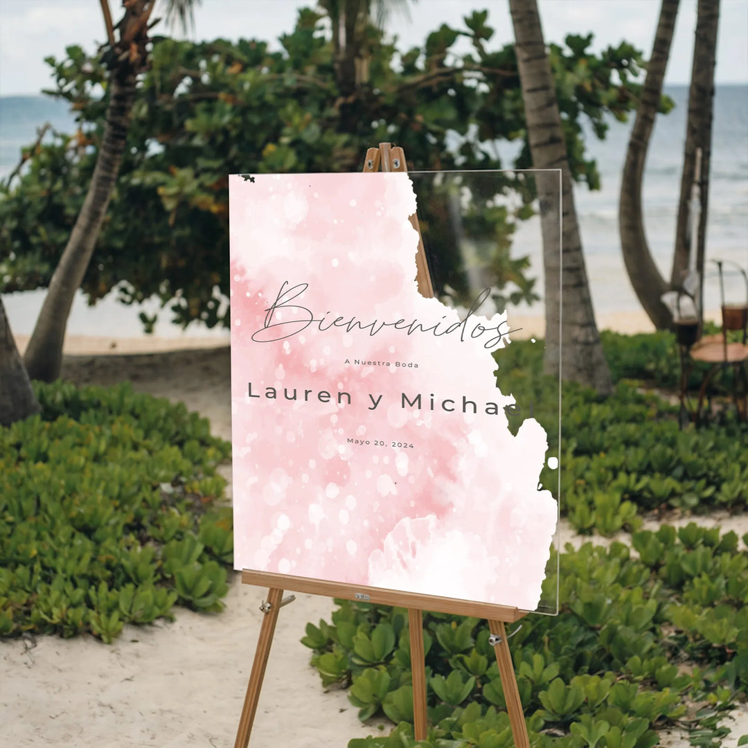 Blush pink acrylic wedding welcome sign with Spanish text on wooden easel at tropical beach