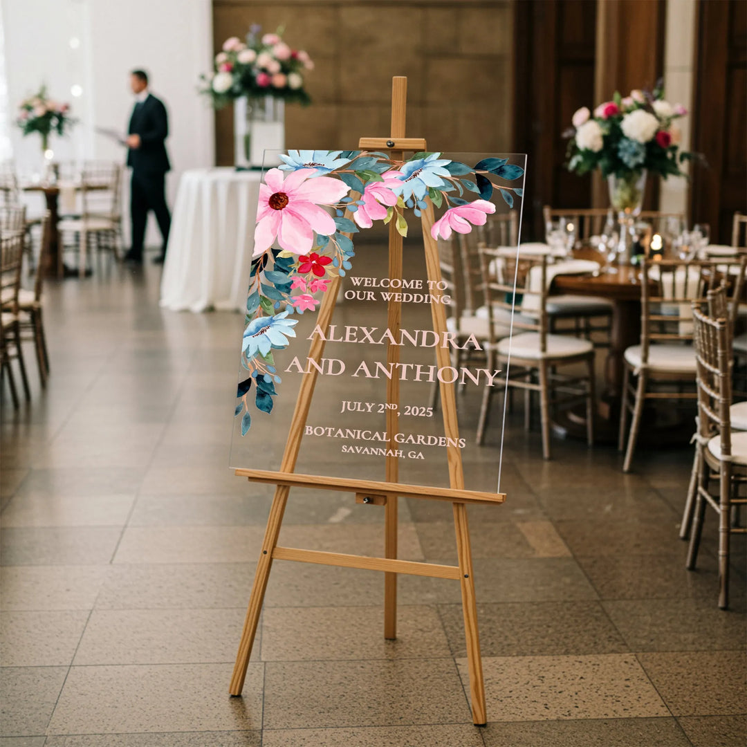 Clear acrylic wedding welcome sign with pink and blue floral design on wooden easel in elegant reception venue