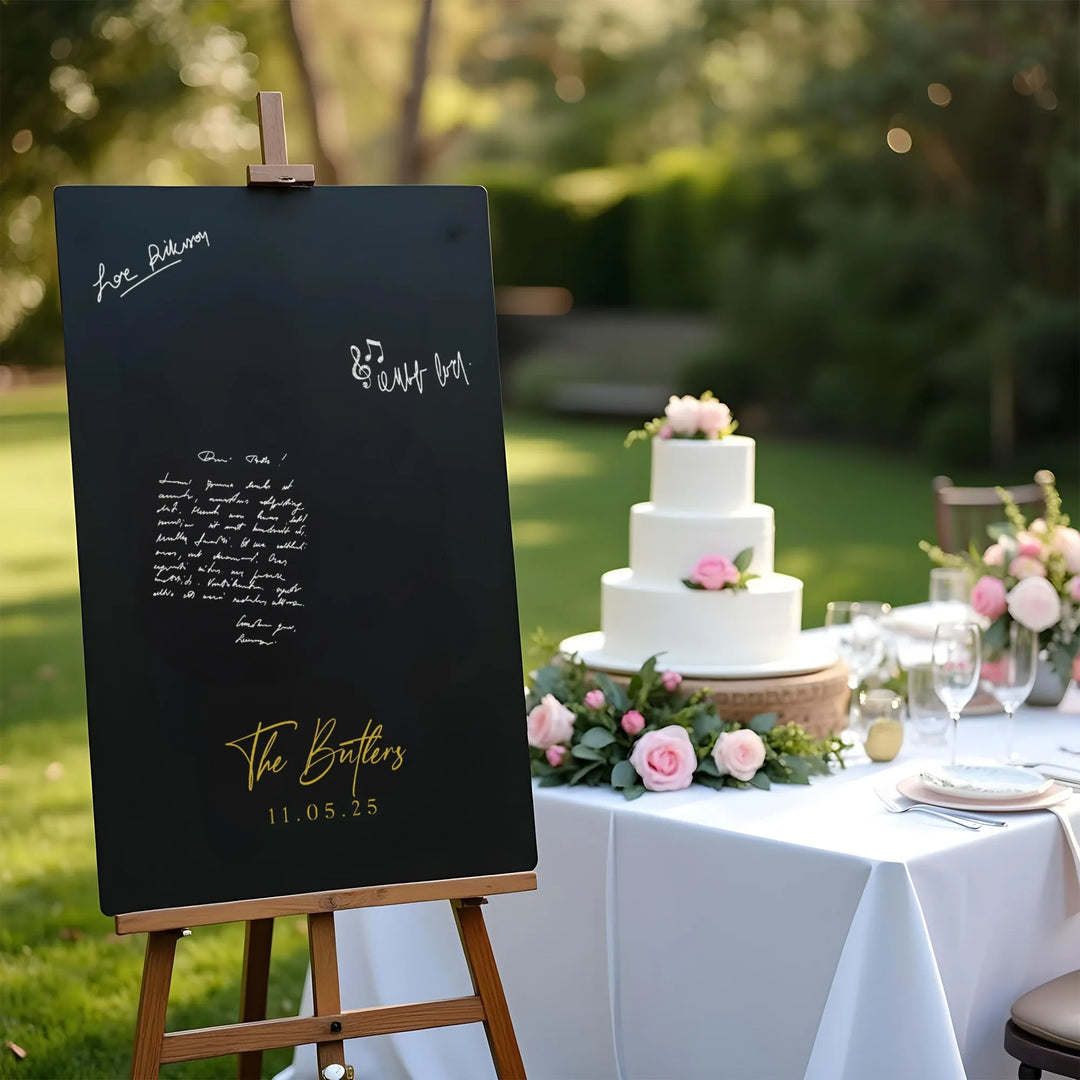 Elegant black wedding guest book sign on easel with gold script The Butlers 11.05.25 beside white tiered cake and pink roses
