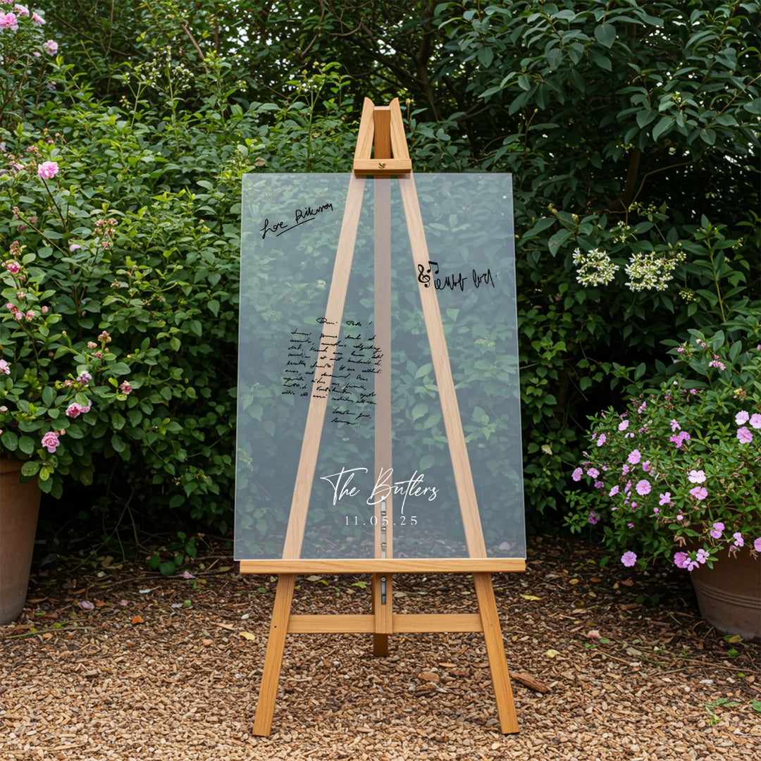 Clear acrylic wedding guest book on wooden easel outdoors with greenery and flowers