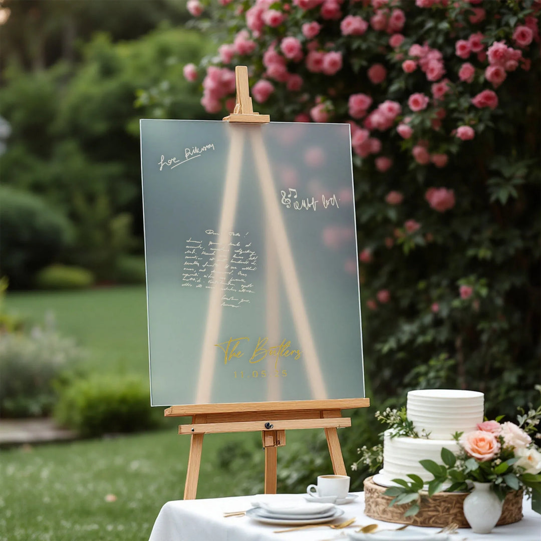 Wedding guest book board with gold lettering on easel next to white tiered cake and floral arrangement in garden