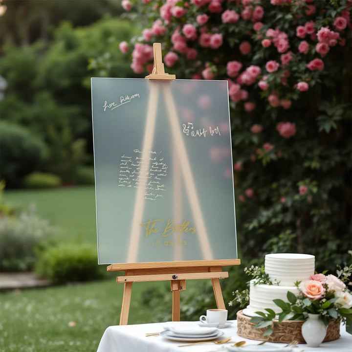Wedding guest book board with gold lettering on easel next to white tiered cake and floral arrangement in garden