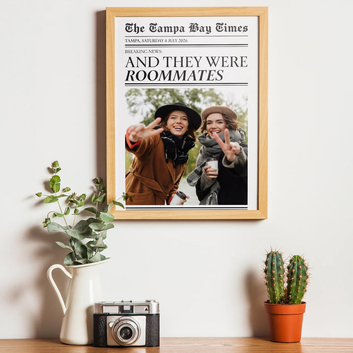 Framed newspaper with two people on a wall above a table with plants and a camera.