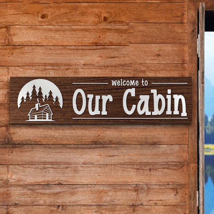 Wooden welcome sign reading Our Cabin with cabin and pine tree silhouette on wood wall