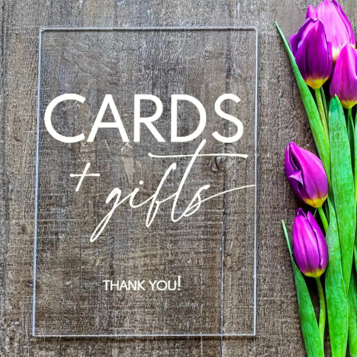 Clear acrylic bridal shower table sign with "Cards + gifts THANK YOU!" text and purple tulips on wooden surface