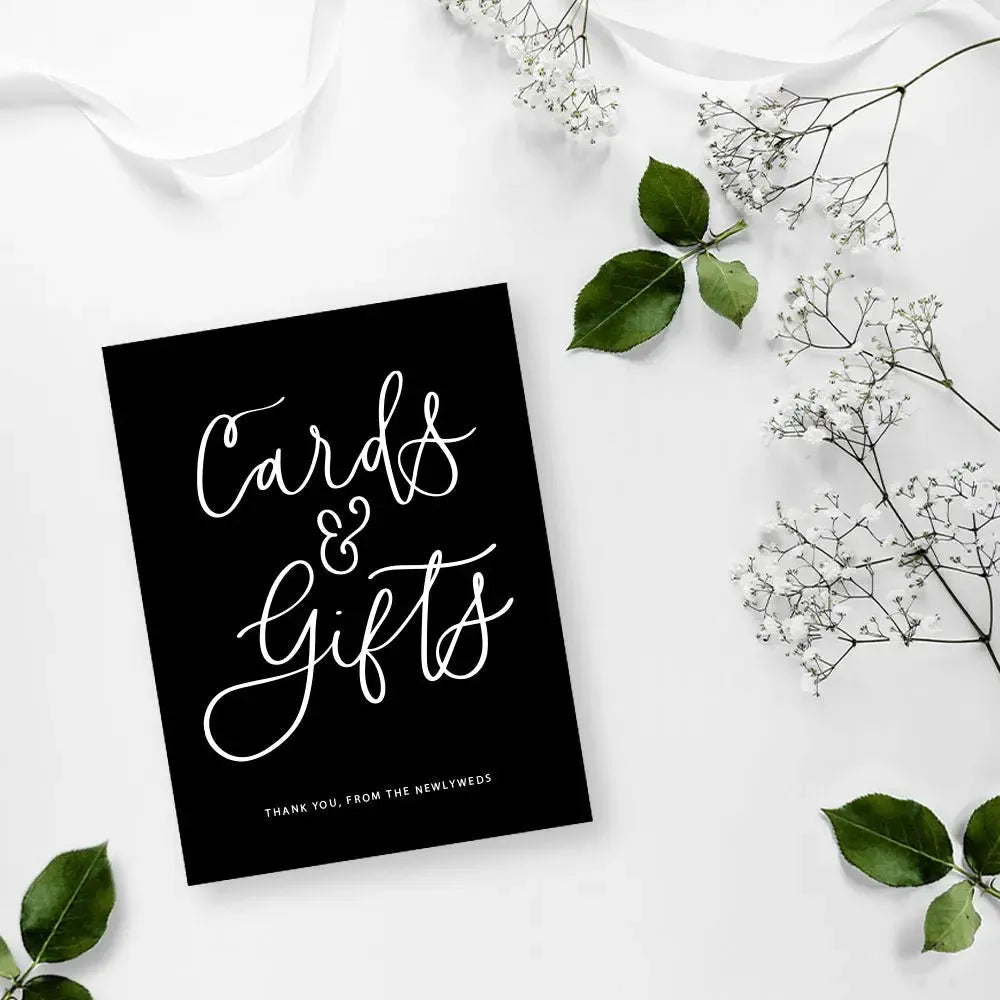 Black wedding table sign reading 'Cards & Gifts' with white flowers and green leaves on white background