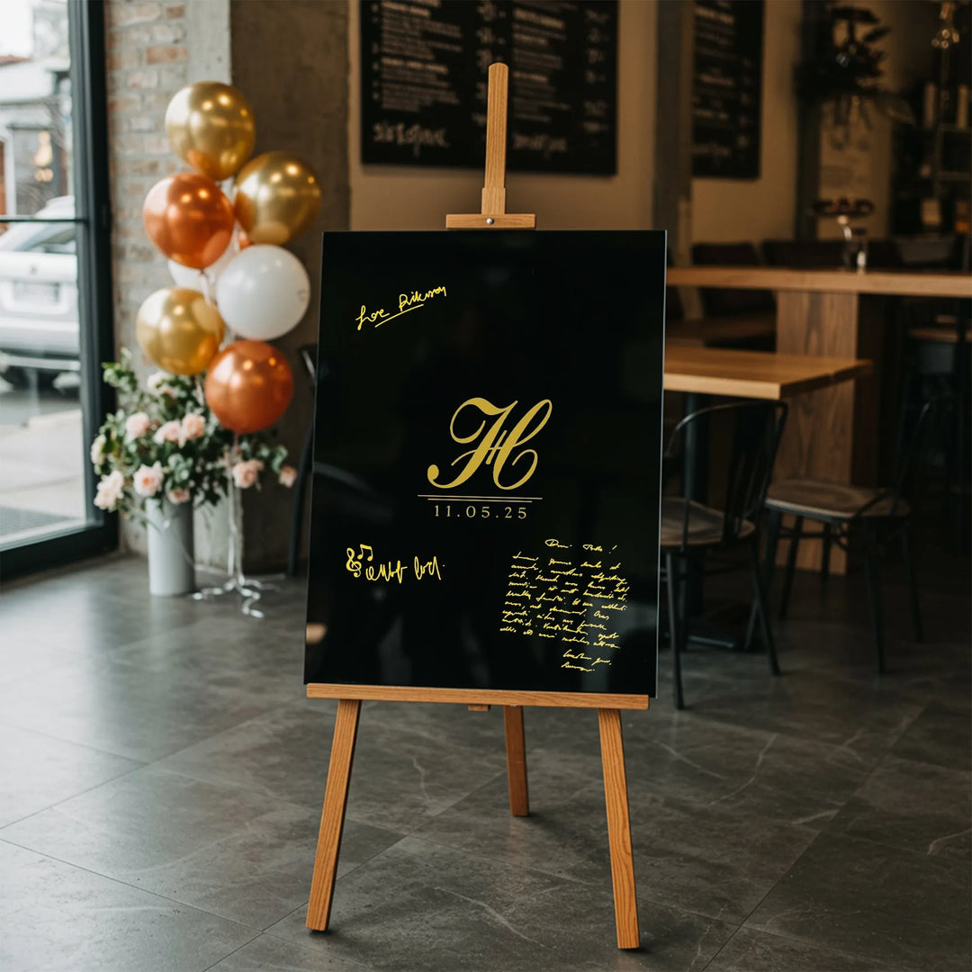 Black wedding guest book sign with gold monogram and date on wooden easel in modern venue with balloons and flowers