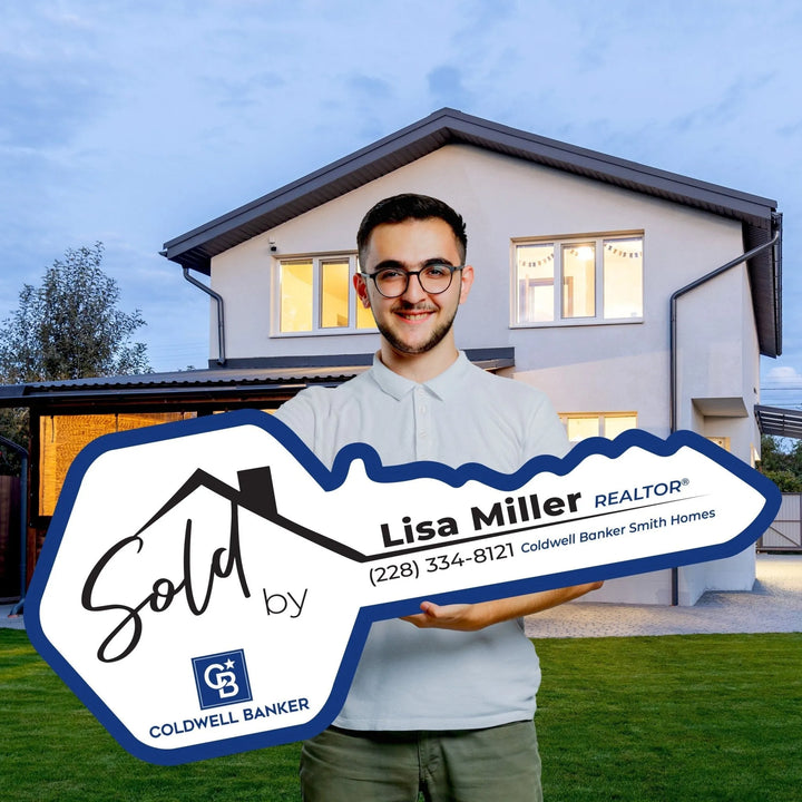 Smiling man holding large Coldwell Banker Sold by Lisa Miller Realtor key sign in front of modern house