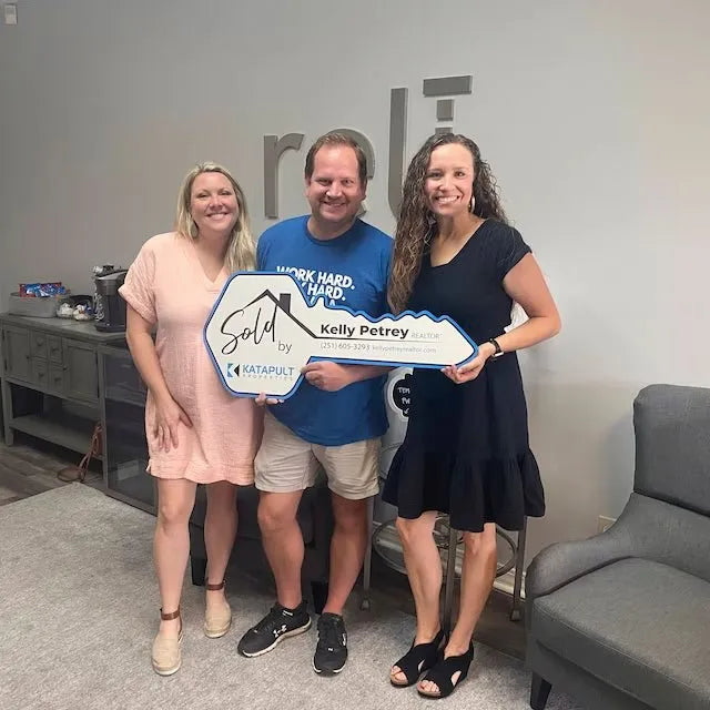 Three smiling people holding a large Sold key sign for Kelly Petrey, Realtor at Katapult Realty, in a modern office