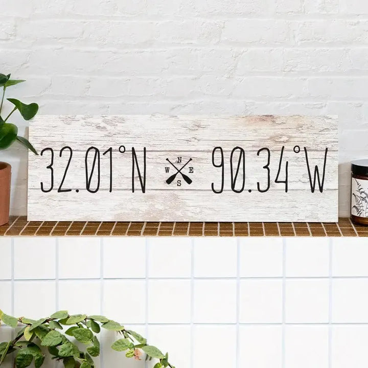 Rustic wood sign with coordinates 32.01°N 90.34°W and compass design on a tiled shelf