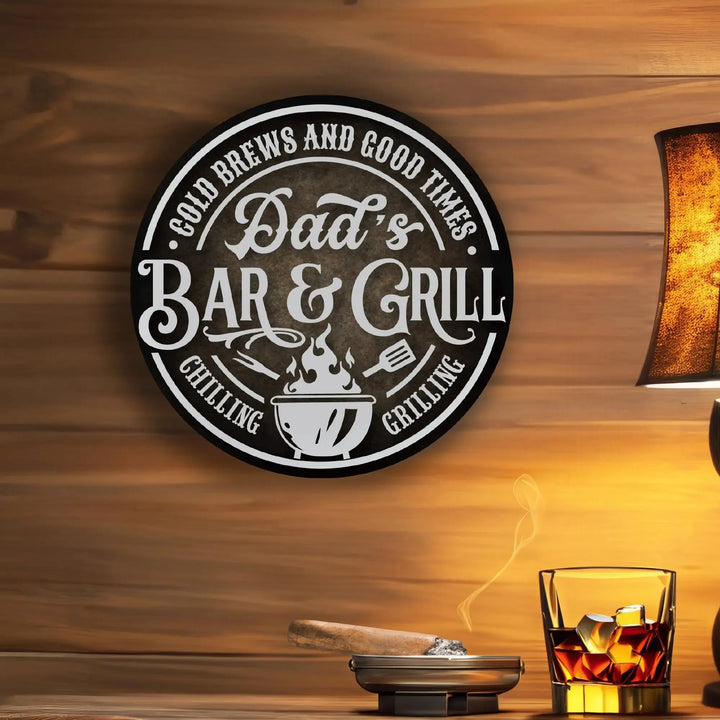 Round metal sign on wood wall reading Dad's Bar & Grill with grilling tools, cigar in ashtray and whiskey glass nearby
