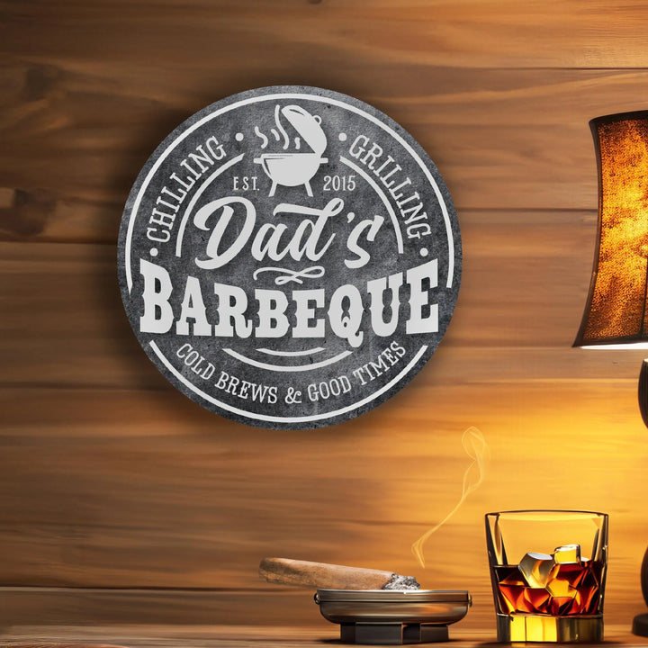 Round metal sign reading Dad's Barbeque Chilling Grilling Cold Brews Good Times on wooden wall