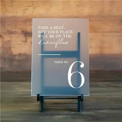 Acrylic wedding table number sign on stand with text about dance floor, rustic wooden background