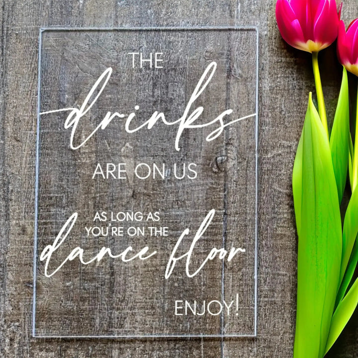 Wedding table sign with text 'The drinks are on us as long as you're on the dance floor' next to pink tulips
