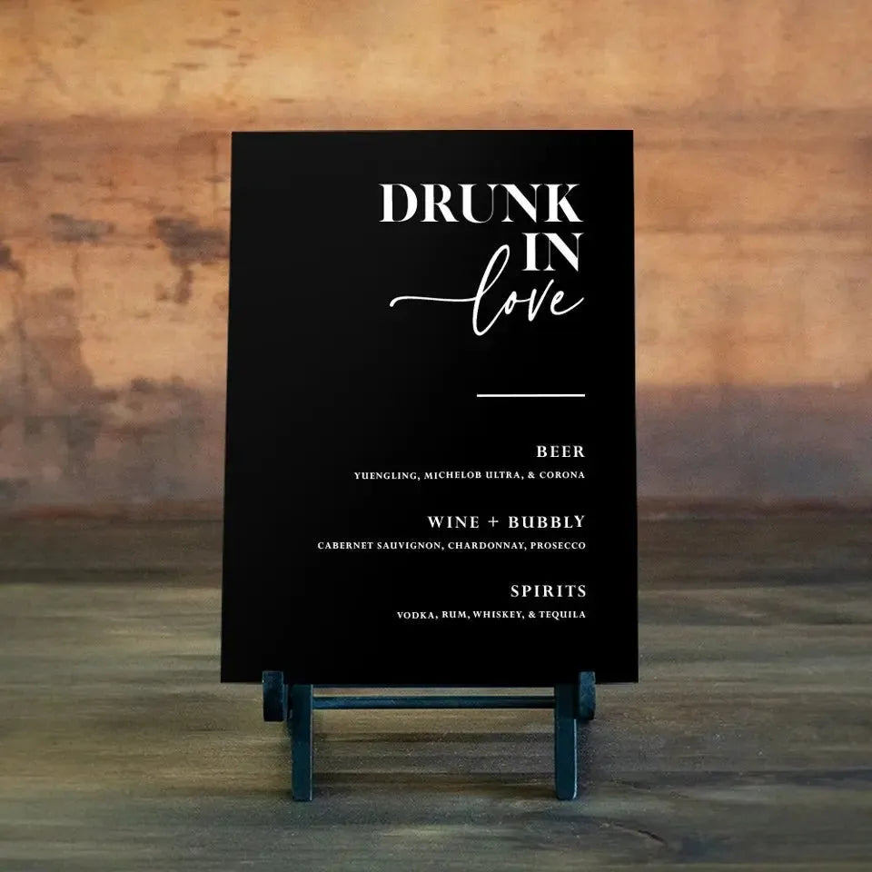 Black wedding bar sign reading 'Drunk in Love' listing beer, wine, bubbly, and spirits on wooden surface