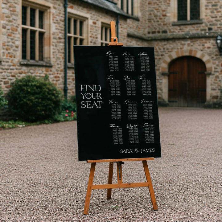 Black wedding seating chart on wooden easel with names list, outdoor stone building background