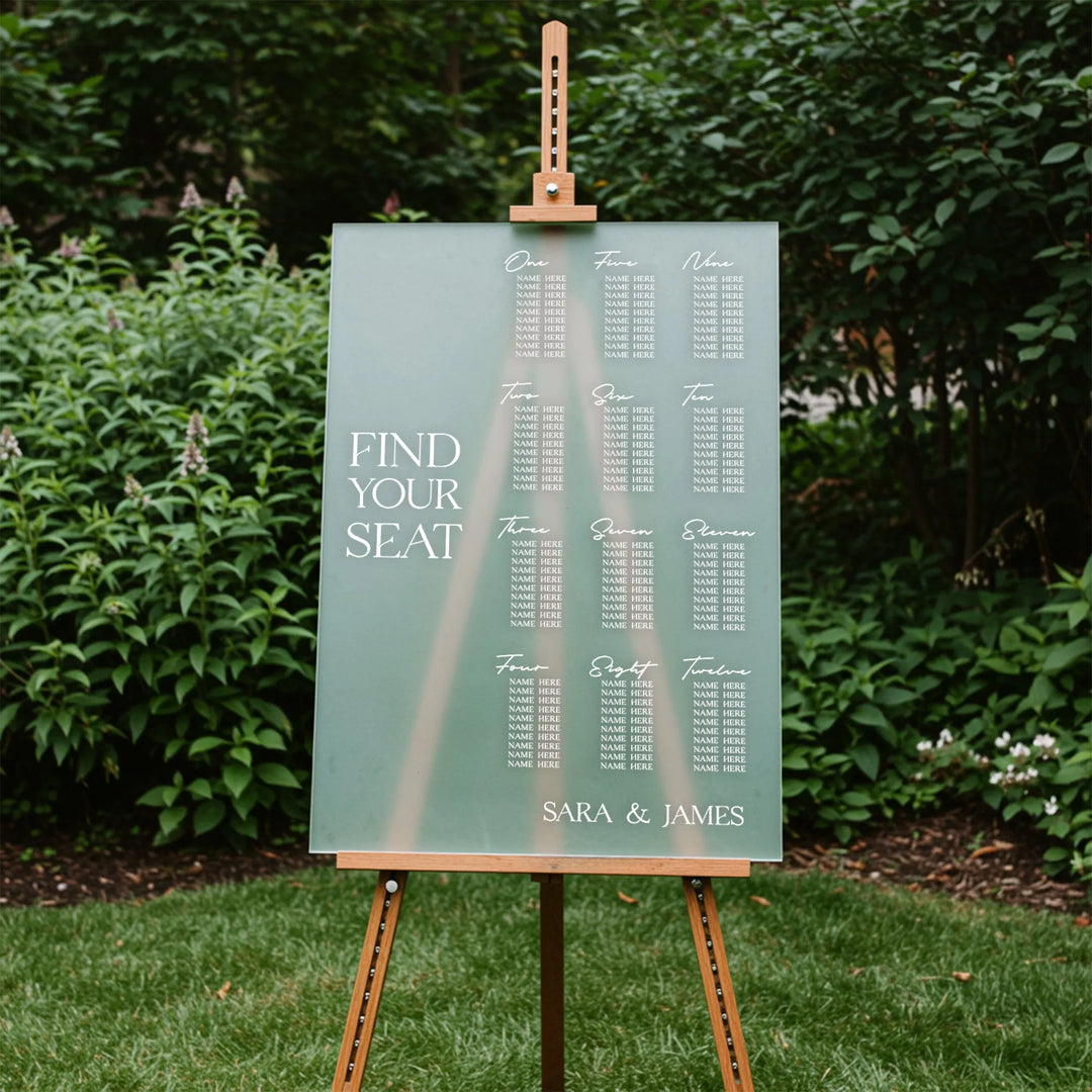 Wedding seating chart on clear acrylic with table numbers and guest names, displayed outdoors on wooden easel