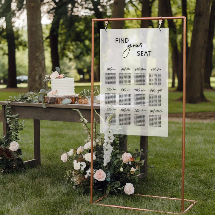 Outdoor wedding seating chart on clear acrylic with copper frame and floral decor near a white wedding cake on wooden table