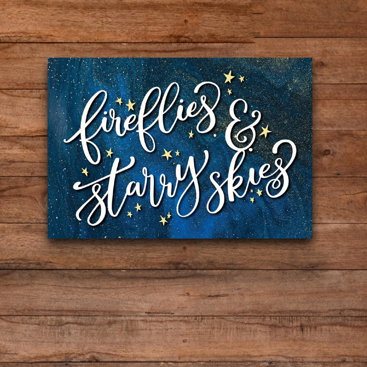 Blue metal wall decor with white script 'fireflies & starry skies' and gold stars on wooden background