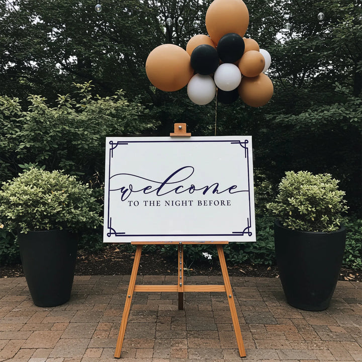Welcome to the Night Before sign on wooden easel with black, white, and gold balloons outdoors
