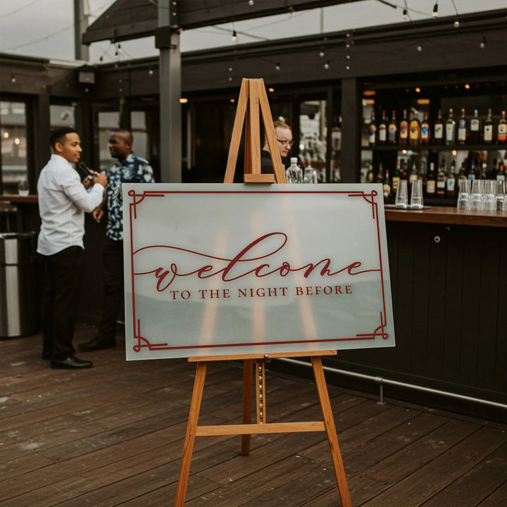 Framed wedding welcome sign on wooden easel reading welcome to the night before at outdoor bar venue