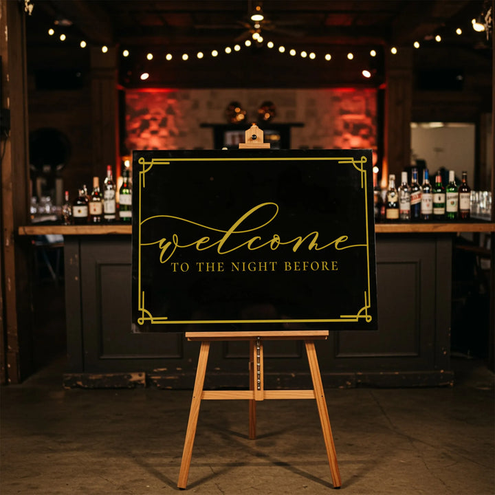 Elegant black and gold wedding welcome sign on wooden easel in dimly lit bar setting