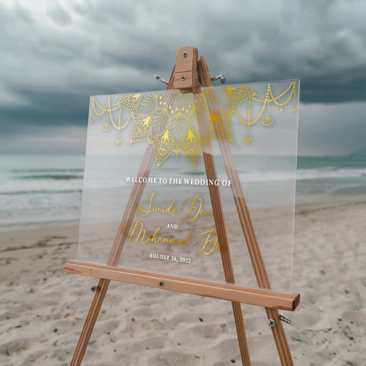 Beach wedding welcome sign on wooden easel with gold mandala design and cloudy sky