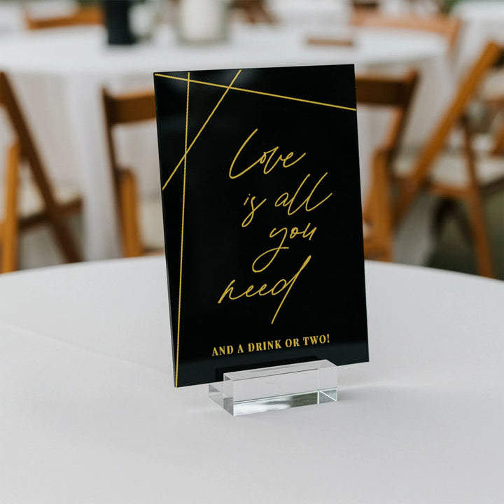 Black geometric wedding bar sign with gold text reading 'Love is all you need and a drink or two' on white table