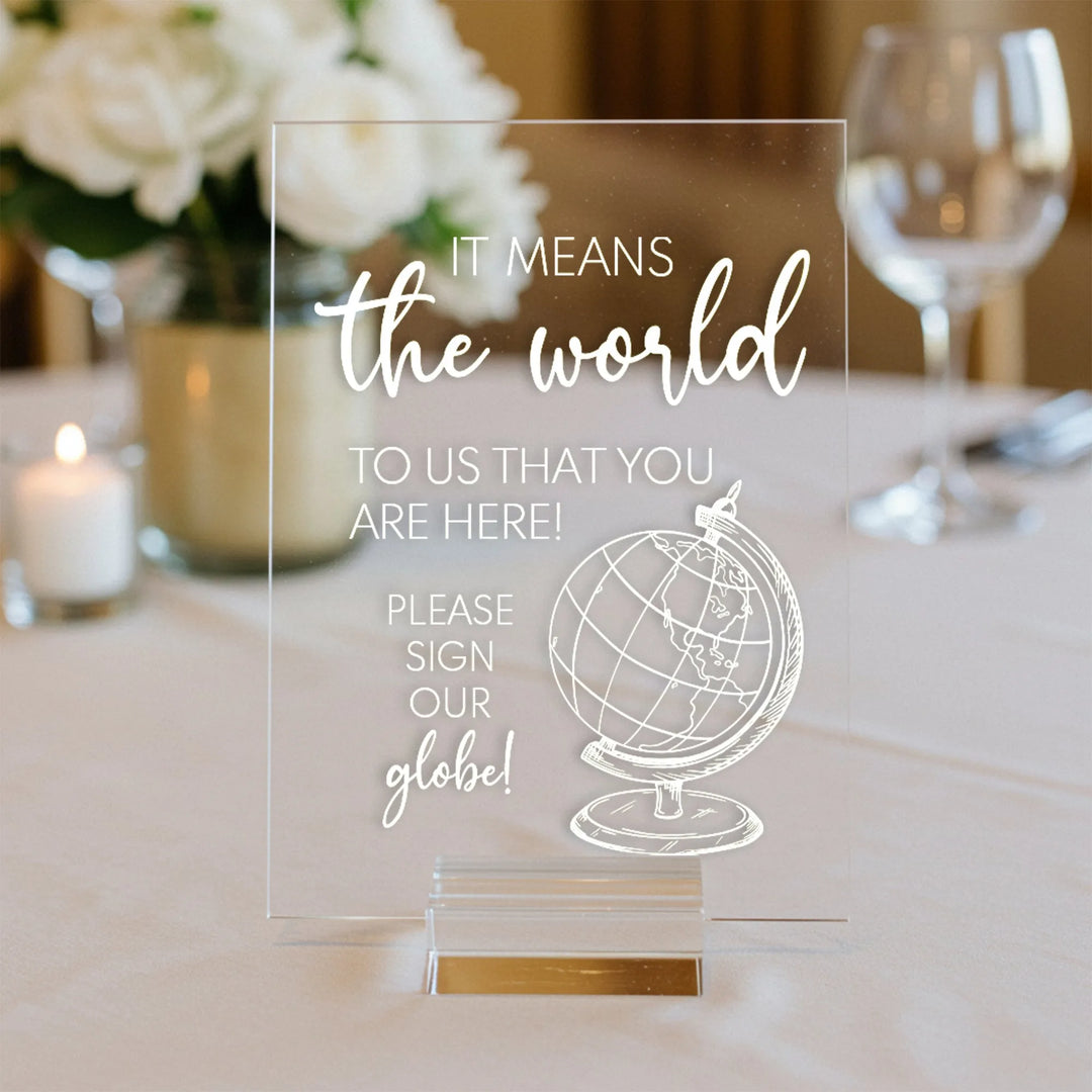 Clear acrylic wedding table sign with white text and globe graphic, white flowers and candle in background