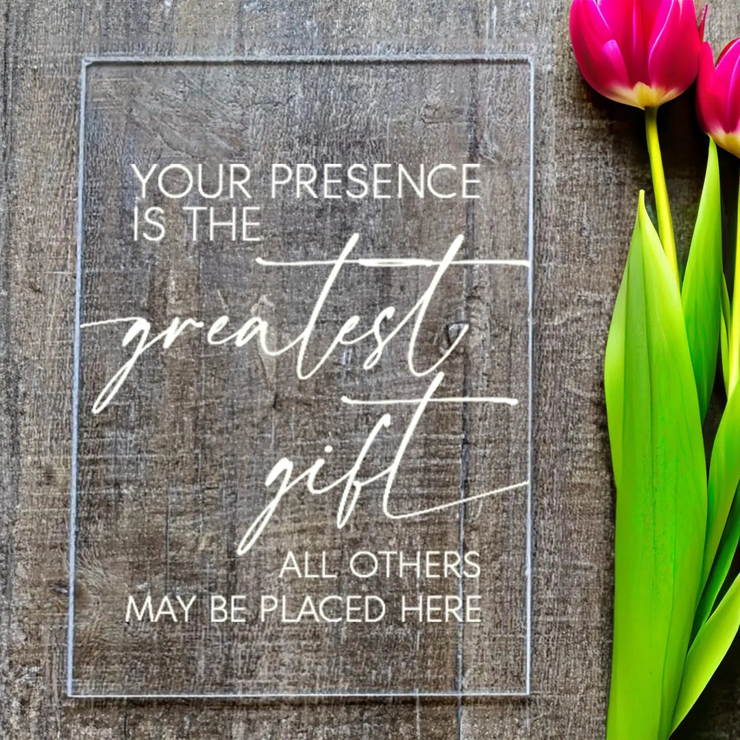 Clear acrylic wedding sign with text Your presence is the greatest gift on wooden surface next to pink tulips