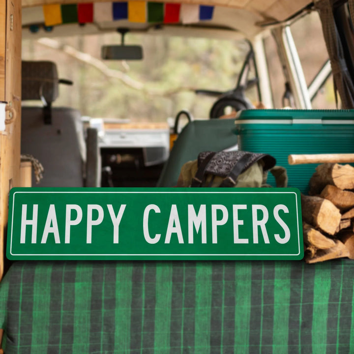 Green metal sign with white text Happy Campers inside a camper van with cooler, backpack, and firewood
