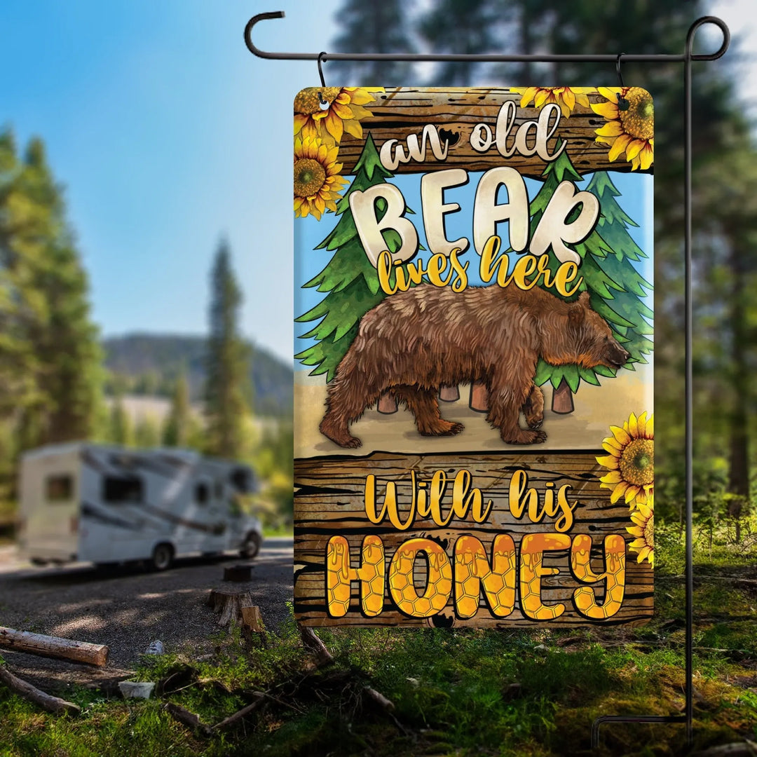 Garden flag with a brown bear walking, pine trees, sunflowers, and text 'an old bear lives here with his honey'