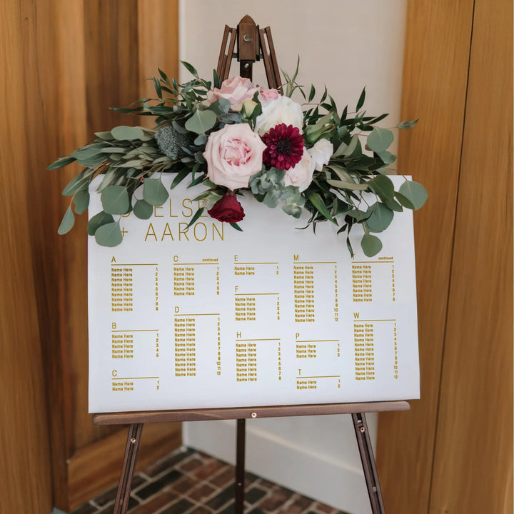 Horizontal wedding seating chart with gold text and floral arrangement on wooden easel